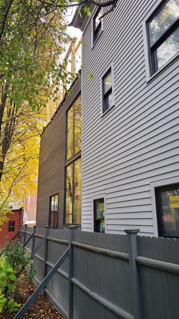 An exterior view showing the connection between the original structure and the modern Hudson House addition. The transition is marked by a tall, vertical glass window assembly that separates the traditional light-grey horizontal siding of the existing townhouse from the dark, contemporary wood-clad volume of the new renovation.