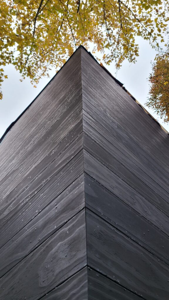 A low-angle detail shot of the Hudson House exterior, focusing on the precision of the mitered corner where two walls of dark, horizontally charred or stained wood siding meet. The natural texture of the wood is visible under the overcast sky, framed by golden autumn leaves at the top of the frame.