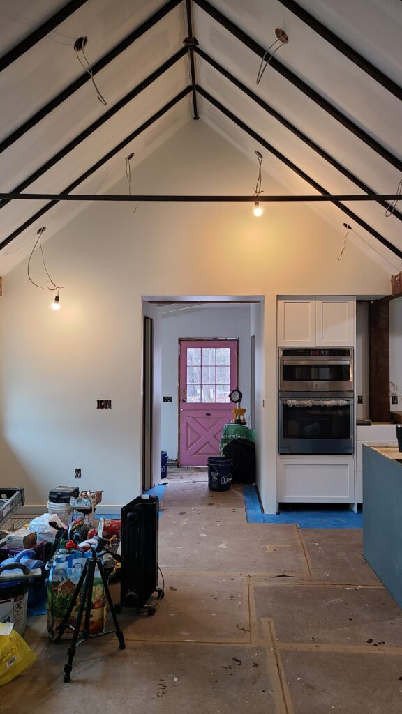 An interior view of the Jardin66 renovation during the construction phase. The kitchen and living area feature a dramatic vaulted ceiling with black structural collar ties. A pink Dutch door serves as a colorful focal point in the background, while white cabinetry and stainless steel appliances are partially installed amid protected subflooring.