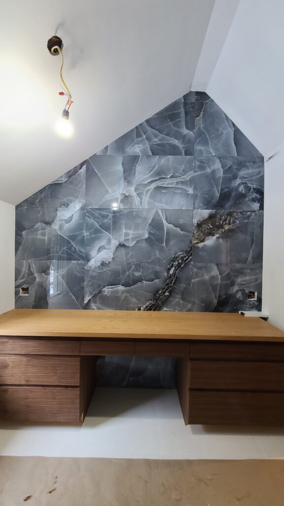 A detailed view of a custom bath vanity area at Jardin66. A rich, dark wood desk with multiple drawers sits beneath a striking wall of book-matched grey marble or stone tiles that follow the line of the sloped ceiling, showcasing high-end interior finishes.