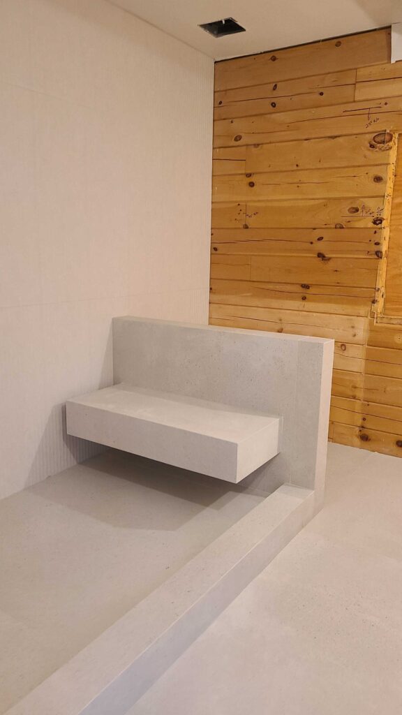 A minimalist, cantilevered concrete bench is integrated into a bathroom or spa area at Olivebridge Reno. The clean lines of the bench contrast with an adjacent rustic wood-plank wall, showcasing the modern-meets-natural aesthetic typical of Hudson Valley modern architecture.