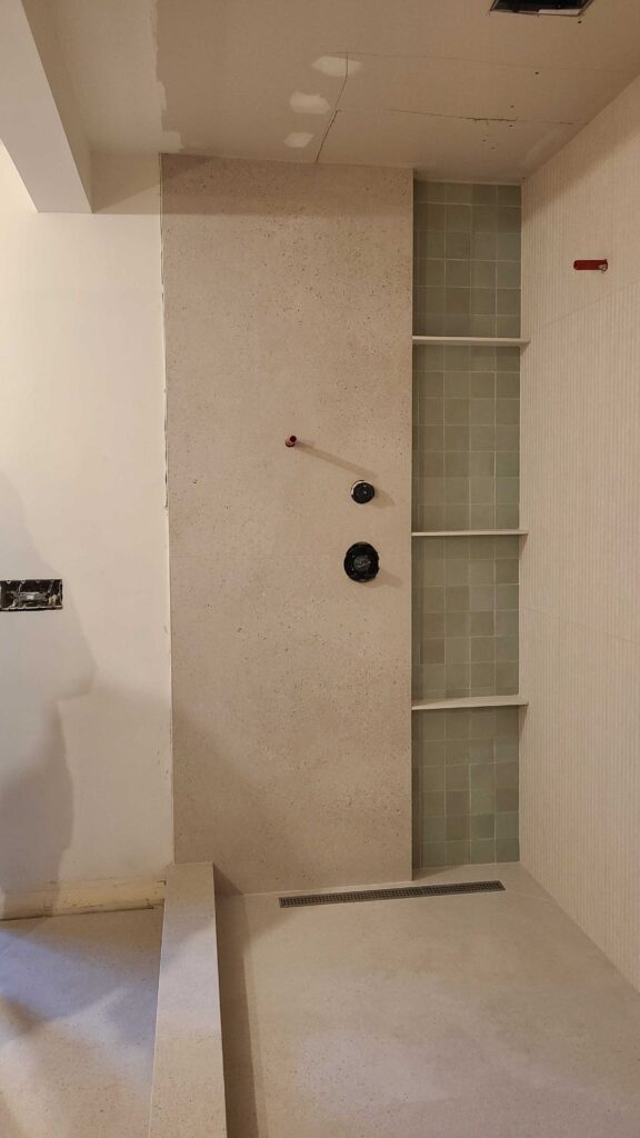 Interior shot of a modern shower installation at Olivebridge Reno. The design features a large, neutral-toned stone slab next to a recessed niche with light green square tiles and built-in shelving. Shower fixtures are positioned on the main slab, illustrating the high-end finishes of this Hudson Valley project.
