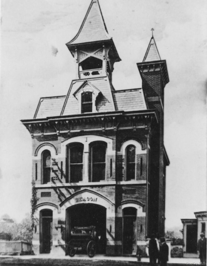 A mid-century black and white photograph of the Beacon Engine Co. No. 1 firehouse. A vintage open-cab fire engine is parked in front. The building shows a two-story brick addition on the left with large rectangular windows, while the original Victorian Gothic structure features arched windows and its signature bell tower.