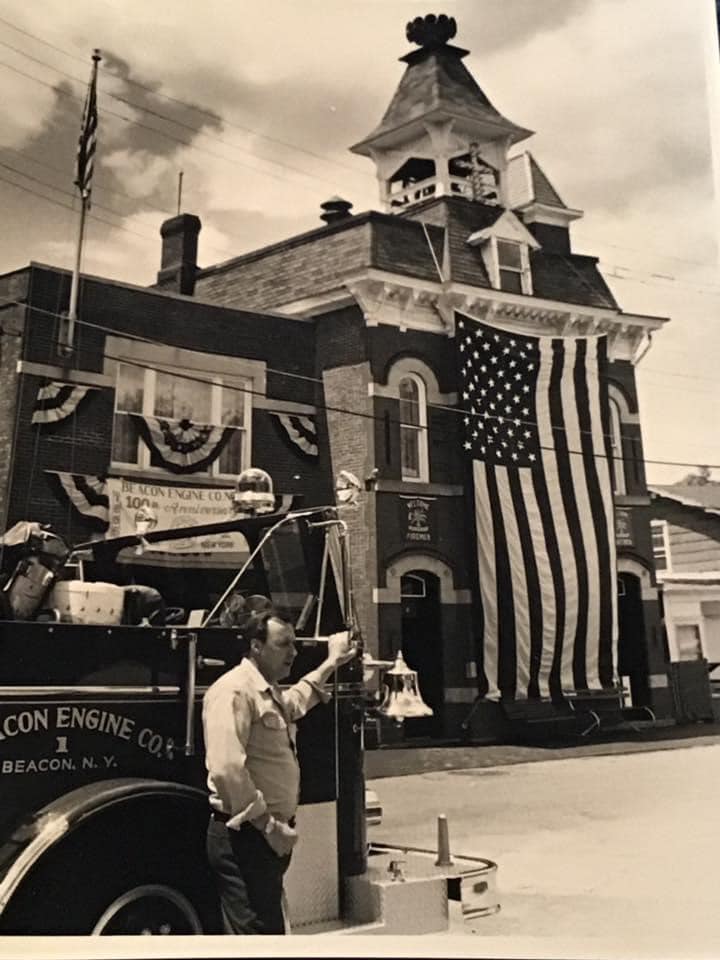 A black and white photo of the firehouse decorated for the Beacon Engine Co. 100th Anniversary. A massive American flag hangs from the front of the brick building, and "100th Anniversary" banners are visible on the left addition. A fireman in uniform stands by a fire truck in the foreground.