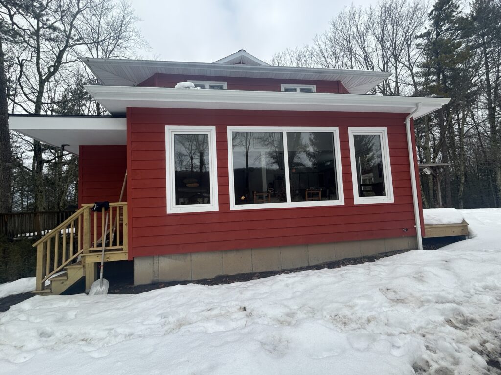 Exterior shot of Accord Addition showing a modern red-sided wing attached to the original home. The addition features a flat roofline with white trim and a trio of large white-framed windows that look out over a snowy landscape. A small wooden staircase with a handrail leads to a side entrance on the left.