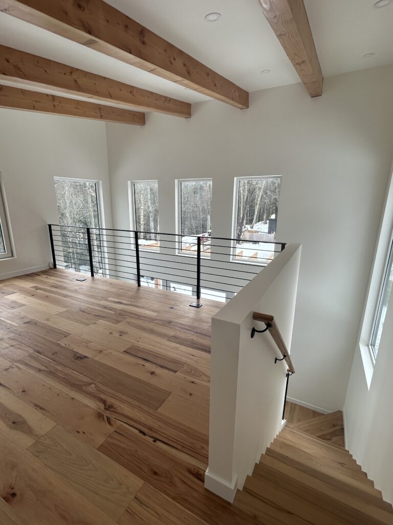 Interior view of the Accord Addition's upper loft, showcasing warm wood plank flooring and exposed timber ceiling beams. A black metal horizontal cable railing lines the staircase and overlooks the double-height space, with natural light pouring in from multiple windows.