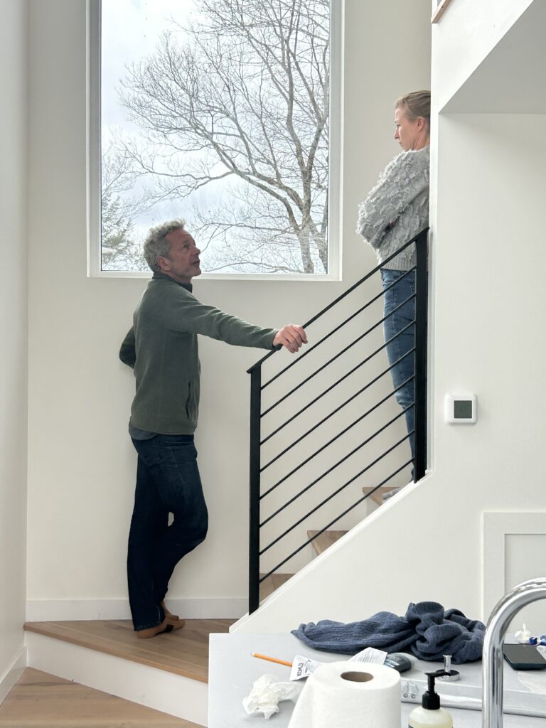 A candid interior shot of the Accord Addition featuring two people standing on a light-wood staircase with a modern black metal railing. Large windows behind them frame the surrounding trees, highlighting the bright, airy atmosphere of the new living space.