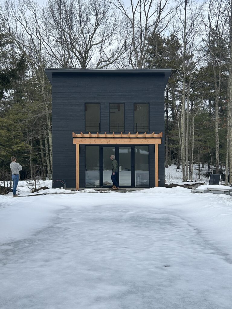 An exterior view of the Accord Addition, a striking two-story modern volume with black horizontal siding. The structure features a wood-beam pergola over a large glass sliding door and three symmetrical windows above, set against a snowy Hudson Valley landscape.