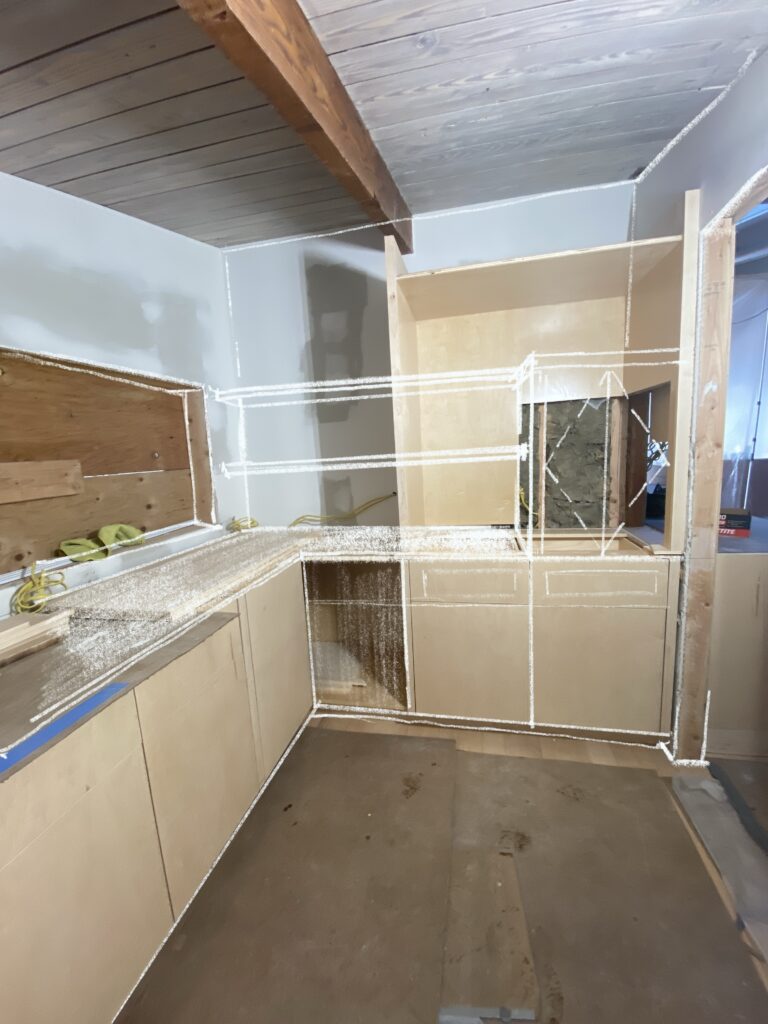 Interior view of the kitchen renovation at MCM Reno, featuring an augmented reality (AR) or digital sketch overlay. The white line drawing visualizes future cabinetry and shelving above the existing light-wood base cabinets, set against a vaulted ceiling with dark wood beams.