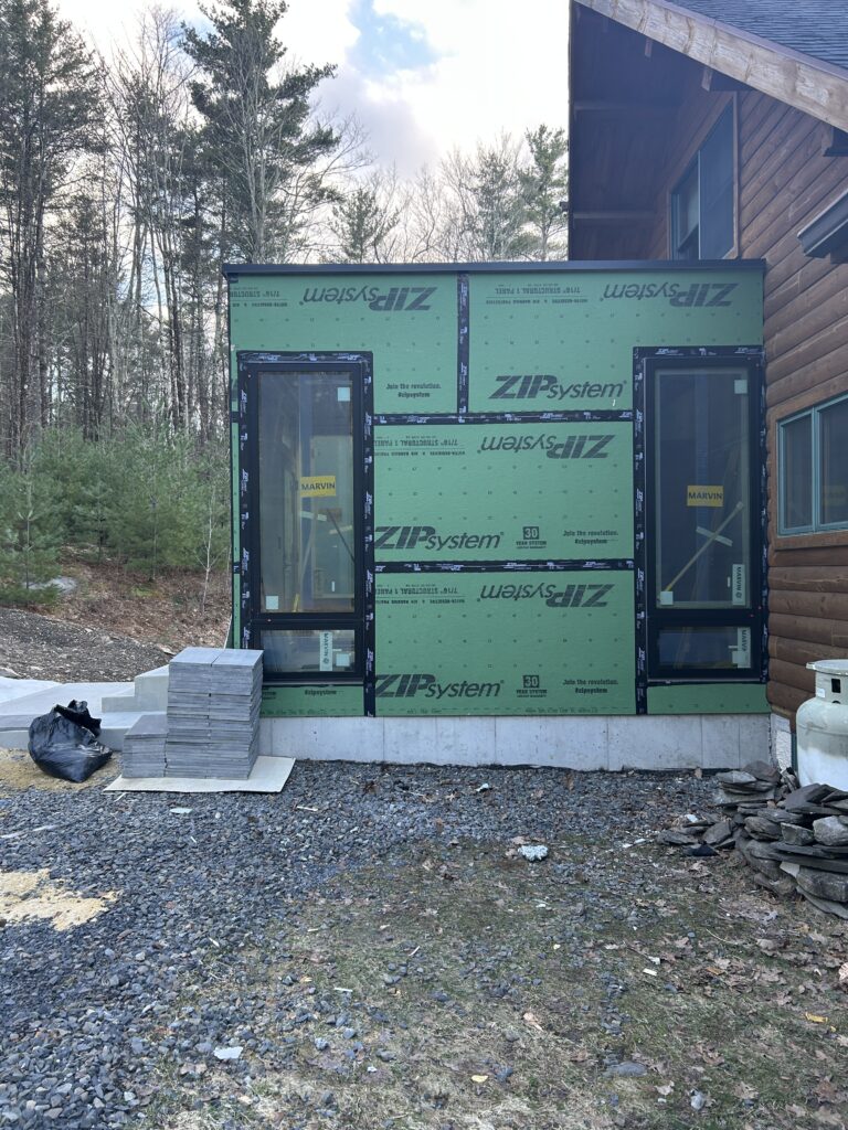 An exterior view of the Olivebridge Reno addition featuring green ZIP System sheathing and two tall, black-framed Marvin windows. The modern structure is attached to a traditional log-style home, with stacks of stone pavers and a gravel workspace in the foreground.