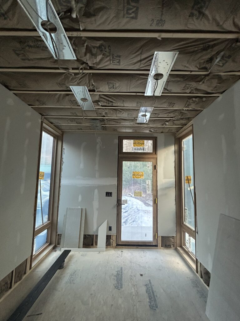 The interior of the Olivebridge Reno addition during the construction phase. The room features exposed ceiling insulation, unfinished drywall, and large windows that fill the space with natural light. A glass-paneled door at the far end offers a view of the surrounding wooded landscape in the Hudson Valley.