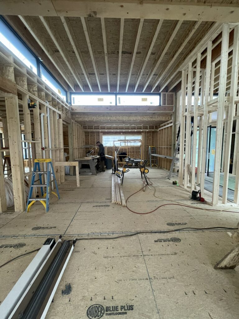 Wide interior view of the open-concept framing at Quattro. Exposed wooden ceiling joists and wall studs create a rhythmic pattern. A row of clerestory windows lets in natural light above the main living area, where construction tools and a miter saw station are set up.