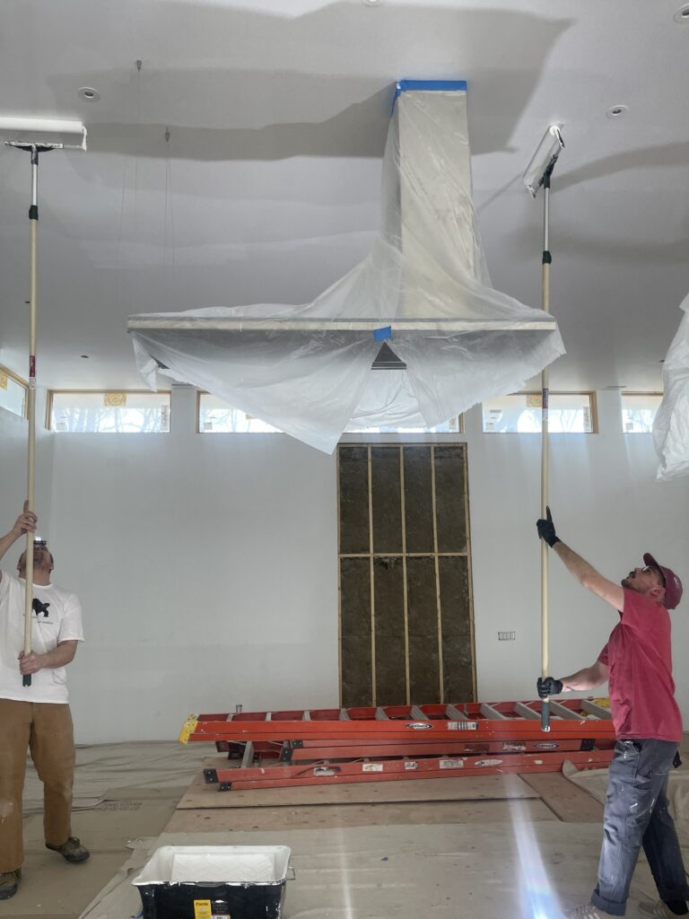 Two builders at Artists Ranch use extension poles to paint the ceiling. The kitchen range hood  is wrapped in protective translucent plastic, with red ladders and construction materials visible in the background of the bright, open space.