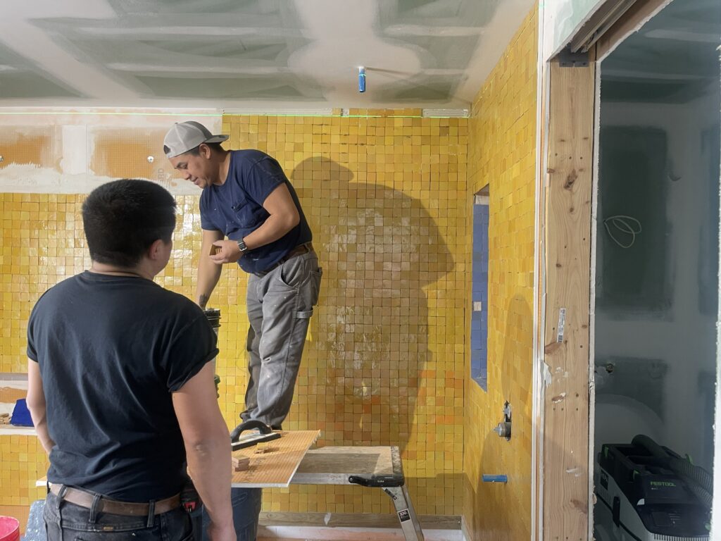 A craftsman installs vibrant yellow Zellige tiles on a bathroom wall at Artists Ranch. Standing on a work platform, the worker carefully aligns the small, hand-finished square tiles against the backdrop of a partially finished drywall ceiling.