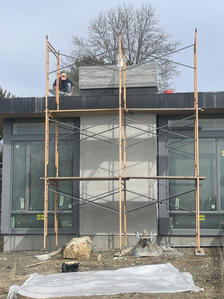 A tall wooden scaffolding tower stands against the exterior of Artists Ranch in the Hudson Valley. A worker is visible on the roofline near a stone-clad chimney feature, with large modern windows and grey siding framing the central structure.
