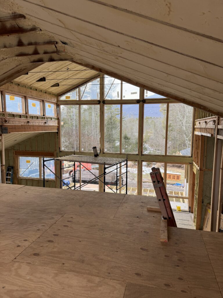 Interior framing of the Pond Lane Modern Renovation showing a double-height living space with a vaulted ceiling. A massive wall of windows is framed out, offering an expansive view of the surrounding trees and hills. Scaffolding and construction materials sit on the plywood subfloor of the upper loft level.