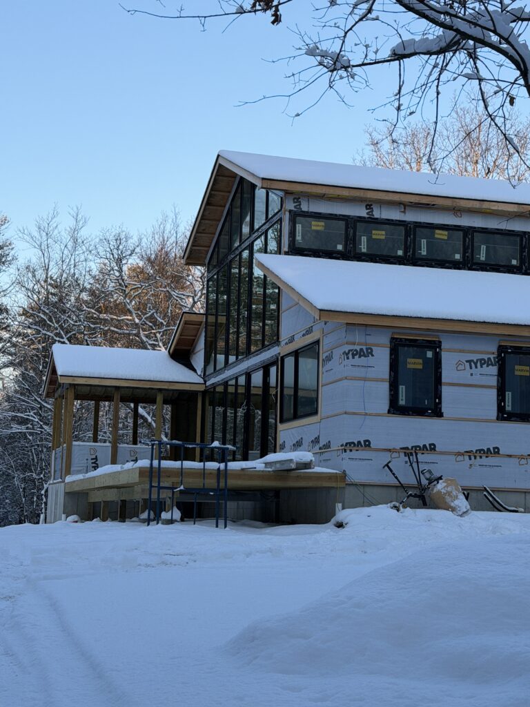 Exterior view of the Pond Lane Modern Renovation during a snowy winter. The home is wrapped in Typar weather barrier with black-framed windows installed. The architectural profile features a dramatic gabled roofline with a large, multi-story glass curtain wall and an integrated porch deck under construction.