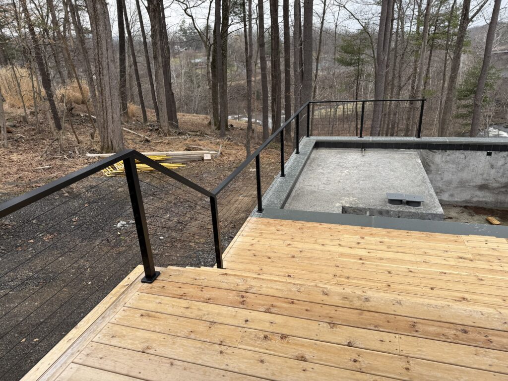 Exterior view of a multi-level wooden deck at Vannstrom featuring a minimalist black metal cable railing. The deck overlooks a sloping wooded site, with a concrete foundation or pool structure visible in the lower section of the construction site.