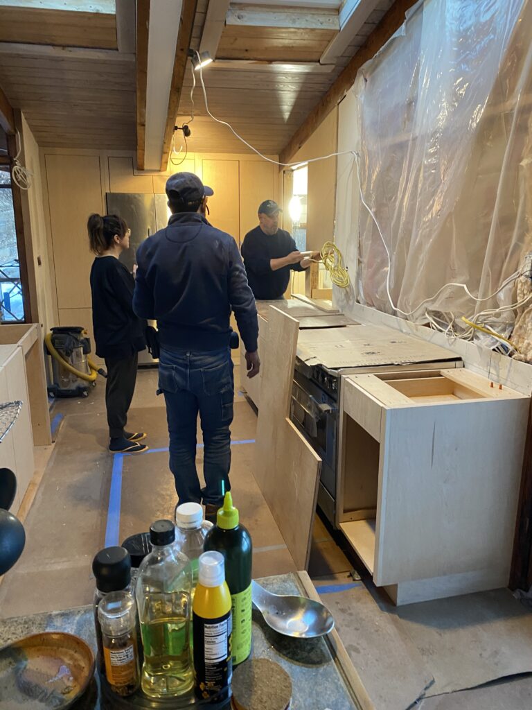 A "behind-the-scenes" look at the kitchen installation at MCM Reno. Three individuals discuss the layout in the active construction zone, which features light-wood cabinetry, blue painter's tape on the floor for templating, and protective plastic over the windows.