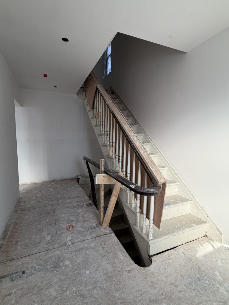 Interior view of the hallway and staircase at Hudson House during renovation. The shot shows a historic wooden staircase with a curved banister and traditional spindles being preserved within a clean, modern white drywall envelope. The subfloor is currently protected with paper, highlighting the blend of historic character and contemporary design.