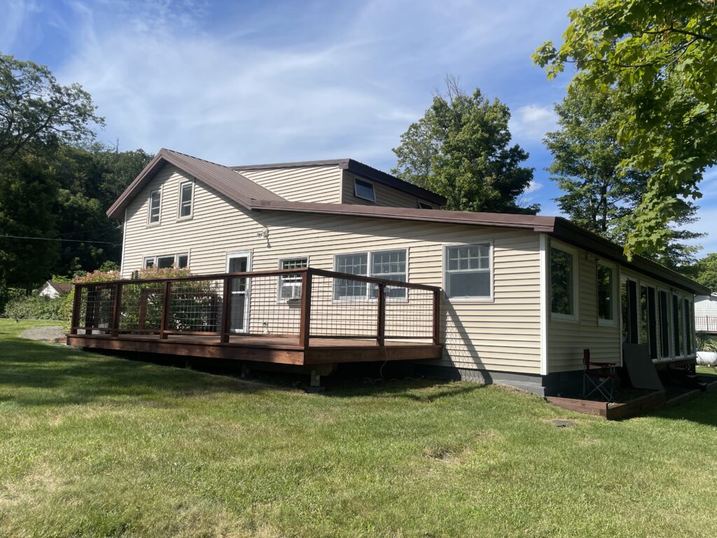 A photograph of the existing exterior of the Acorn Hill residence in Olivebridge, NY. The house is a multi-level structure with tan vinyl siding, a brown metal roof, and a large wooden deck featuring a wire mesh railing. The surrounding landscape is a lush green lawn with mature trees under a blue sky.