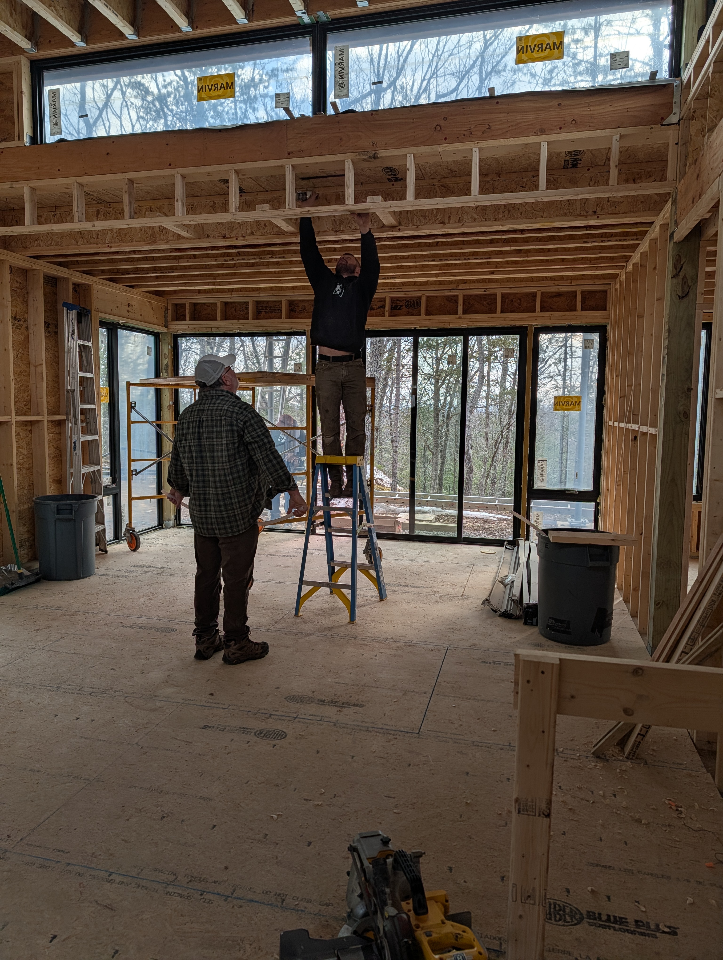 Interior view of the framing phase at Quattro, showing two builders working near a large window wall. One worker stands on a blue stepladder reaching toward the ceiling joists, while the other observes from the subfloor. Marvin windows and tall glass doors are visible in the background.