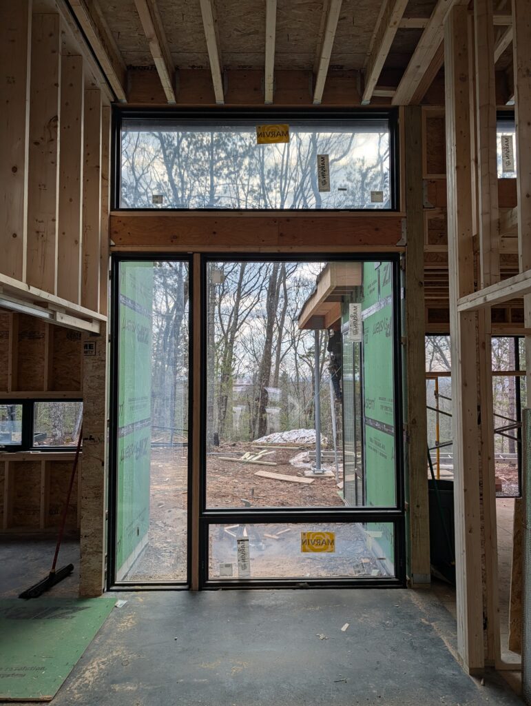 Interior perspective looking out through a large, black-framed Marvin window and clerestory assembly at Quattro. The view through the glass shows the wooded Saugerties landscape and the exterior green sheathing of an adjacent wing of the home.