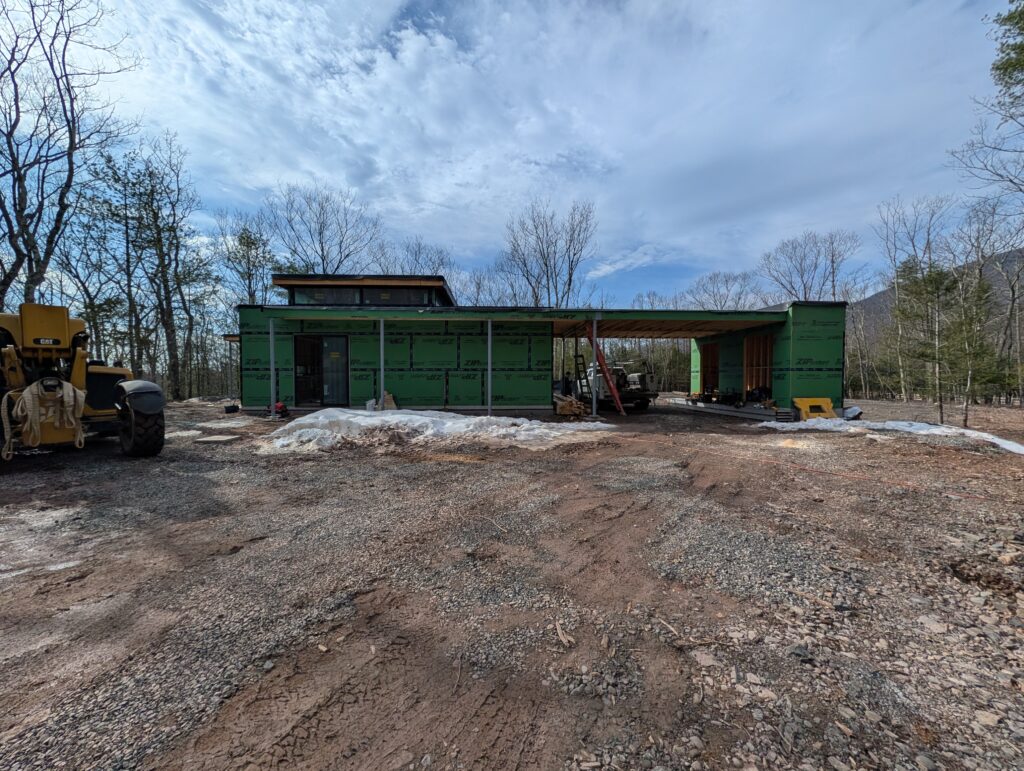 Exterior wide shot of Quattro, a custom modern home under construction in Saugerties. The structure features green ZIP System sheathing, a flat roofline with a clerestory level, and an integrated carport. The surrounding site is a dirt and gravel clearing with bare trees in the background.