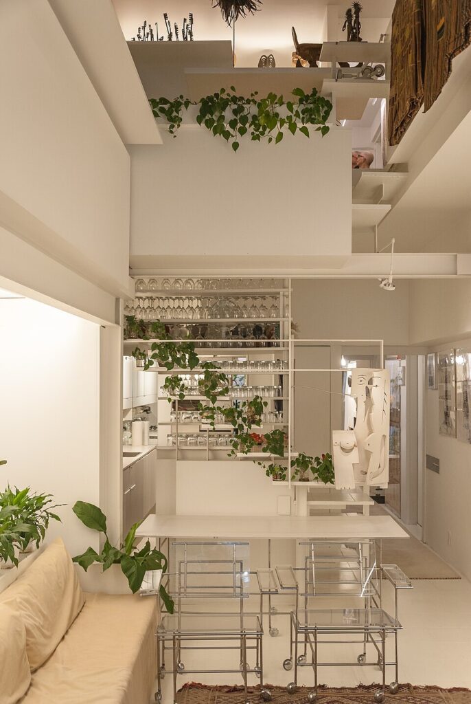 Modern minimalist apartment interior showing vertical spatial layering. Features include white-painted structural beams, integrated plant life, and tiered shelving that creates a sense of depth and openness.