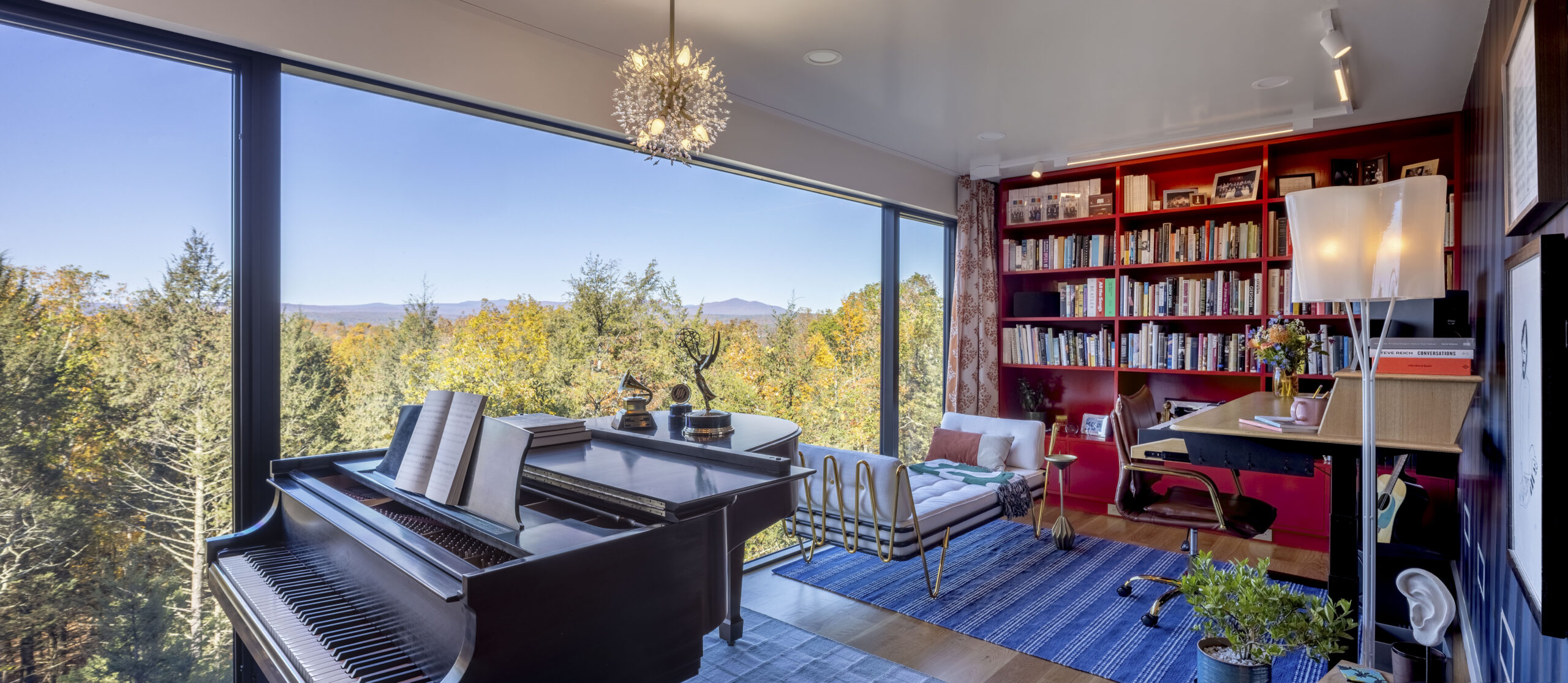 A sun-drenched music studio dominated by a floor-to-ceiling window overlooking the forest. A black grand piano sits in the foreground. Against the far wall, bright red built-in bookshelves house a large collection, and a modern desk is paired with a brown leather chair on a blue striped rug.