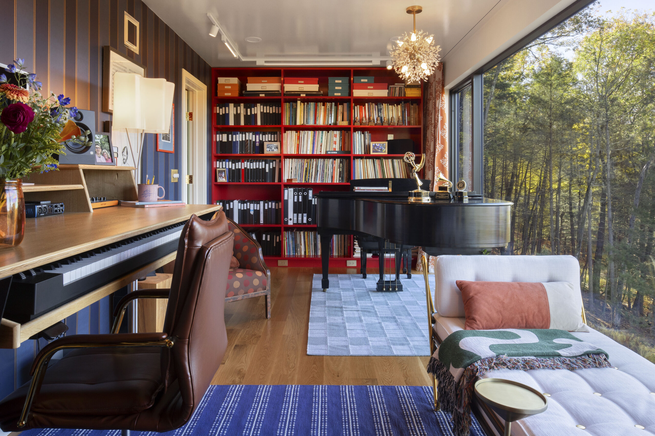An alternate angle of the music studio, showing a close-up of the workstation with an integrated keyboard. A white daybed with an orange accent pillow sits by the window, offering a place to rest while overlooking the woods. The room features a dark-striped feature wall and a sparkling dandelion-style chandelier.