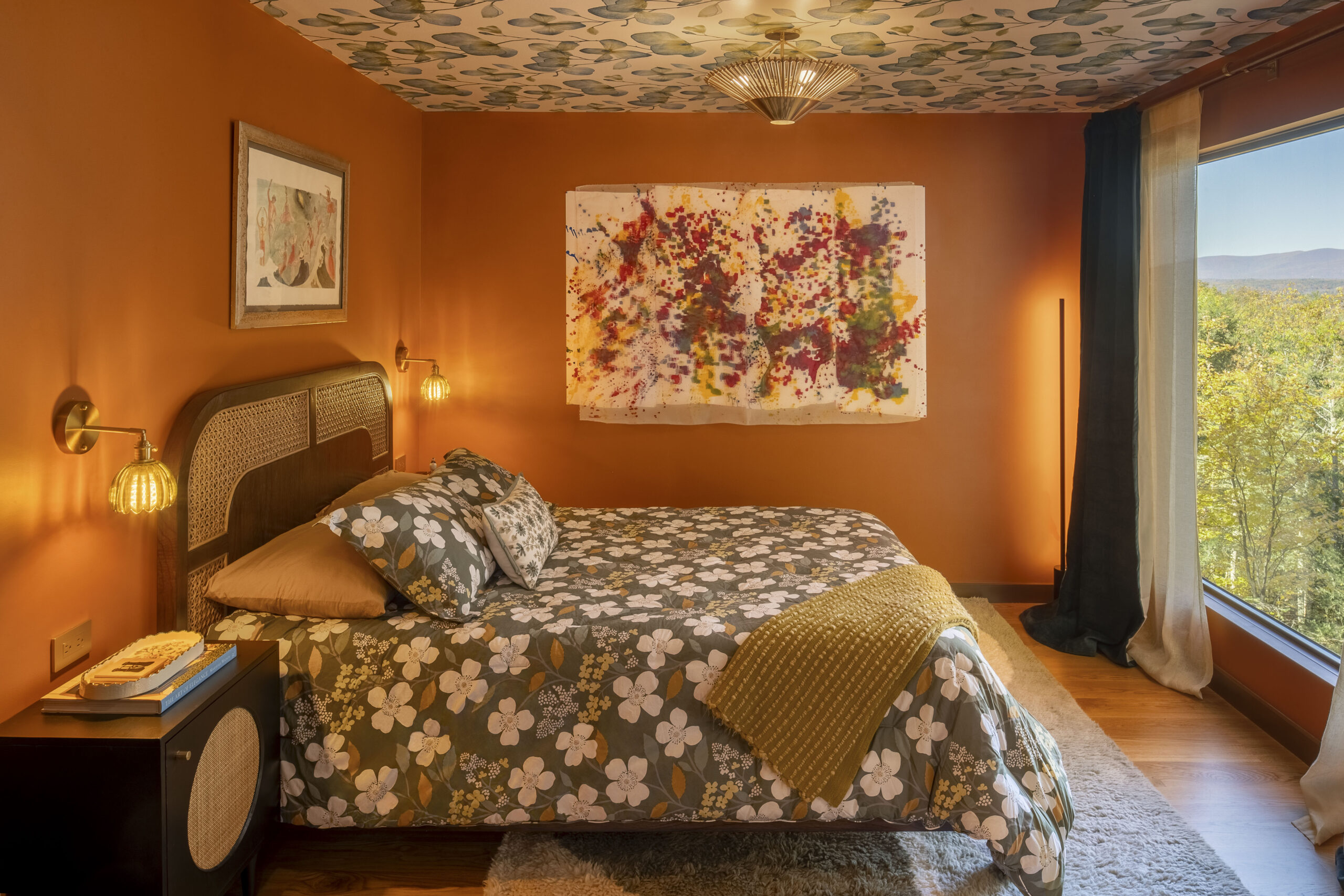 A warm, inviting bedroom with rich terracotta-orange walls and a custom-patterned ceiling featuring blue and green leaves. The bed has a dark wood headboard with cane inserts and is covered in a green floral duvet. A large, abstract splatter-paint canvas hangs above the bed, and a wide window provides a panoramic view of the Hudson Valley autumn foliage.