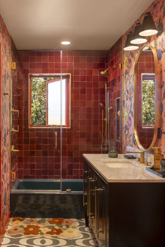A high-contrast bathroom featuring deep crimson square tiles in the walk-in shower and a dark teal bathtub. The vanity wall is covered in a bold pink and blue floral wallpaper. A marble-topped dark wood vanity with gold hardware is paired with a gold-rimmed oval mirror and black conical sconces.