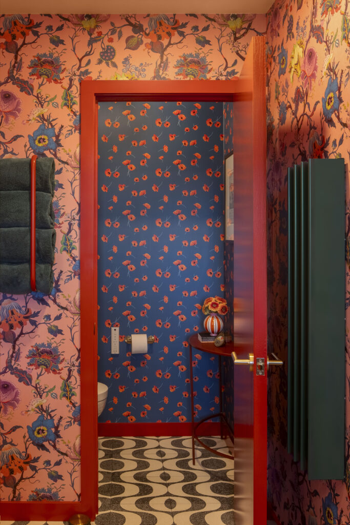 A view into a powder room showing a fearless mix of patterns. The outer walls feature a large-scale pink and blue botanical print, while an open bright-red door reveals an inner room with a deep blue poppy-patterned wallpaper. The floor is a monochromatic black and white retro-wave tile.