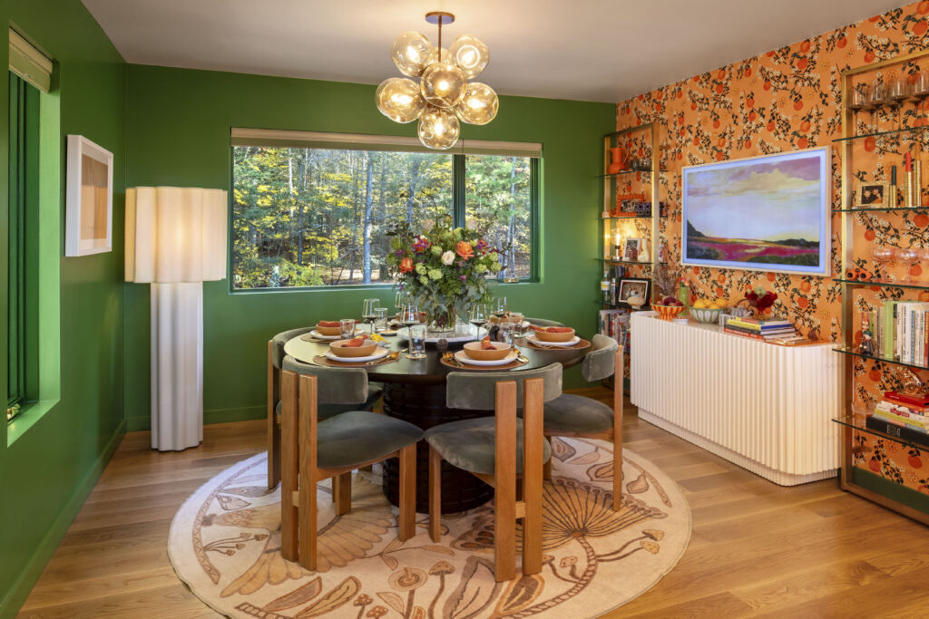 A bold dining room with forest-green walls and a feature wall covered in a vintage orange citrus-patterned wallpaper. A round black dining table is surrounded by grey velvet chairs on a whimsical mushroom-patterned rug. A white fluted sideboard and a large glass-globe chandelier complete the high-energy, eclectic design.