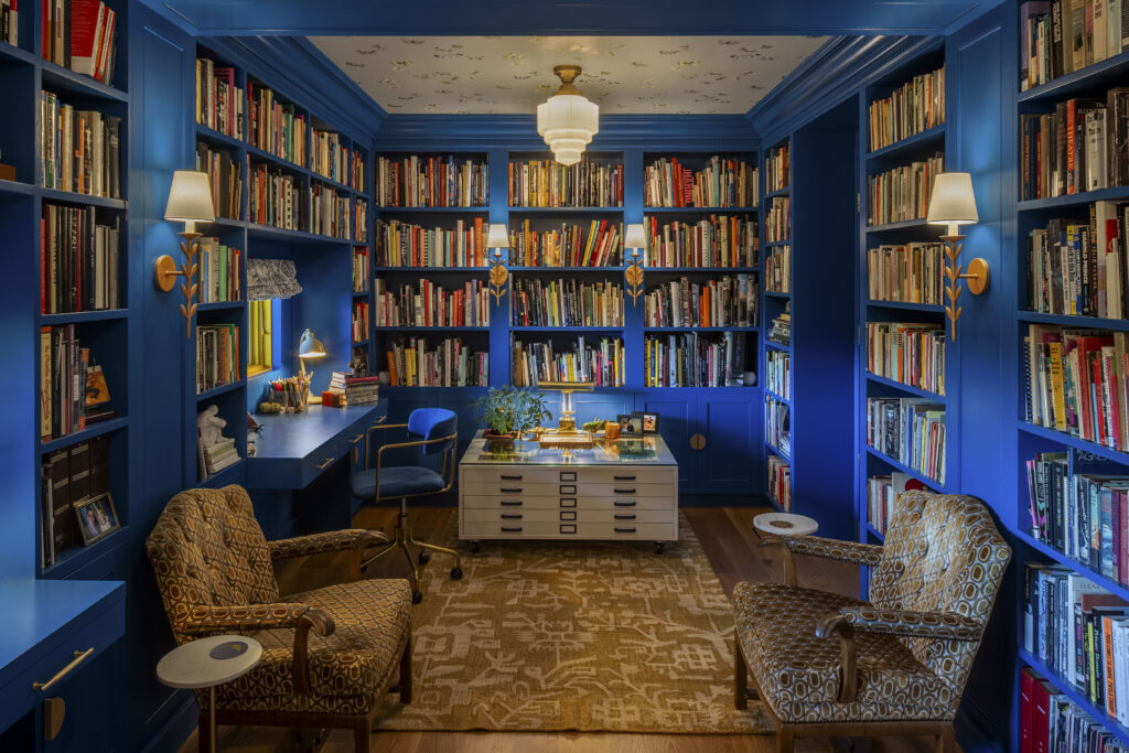 A stunning library where the walls, ceiling, built-in bookshelves, and desk are all painted the same vibrant shade of royal blue. The room is filled with hundreds of books. Two patterned armchairs face each other over a tan patterned rug, with a large white map cabinet used as a central table.