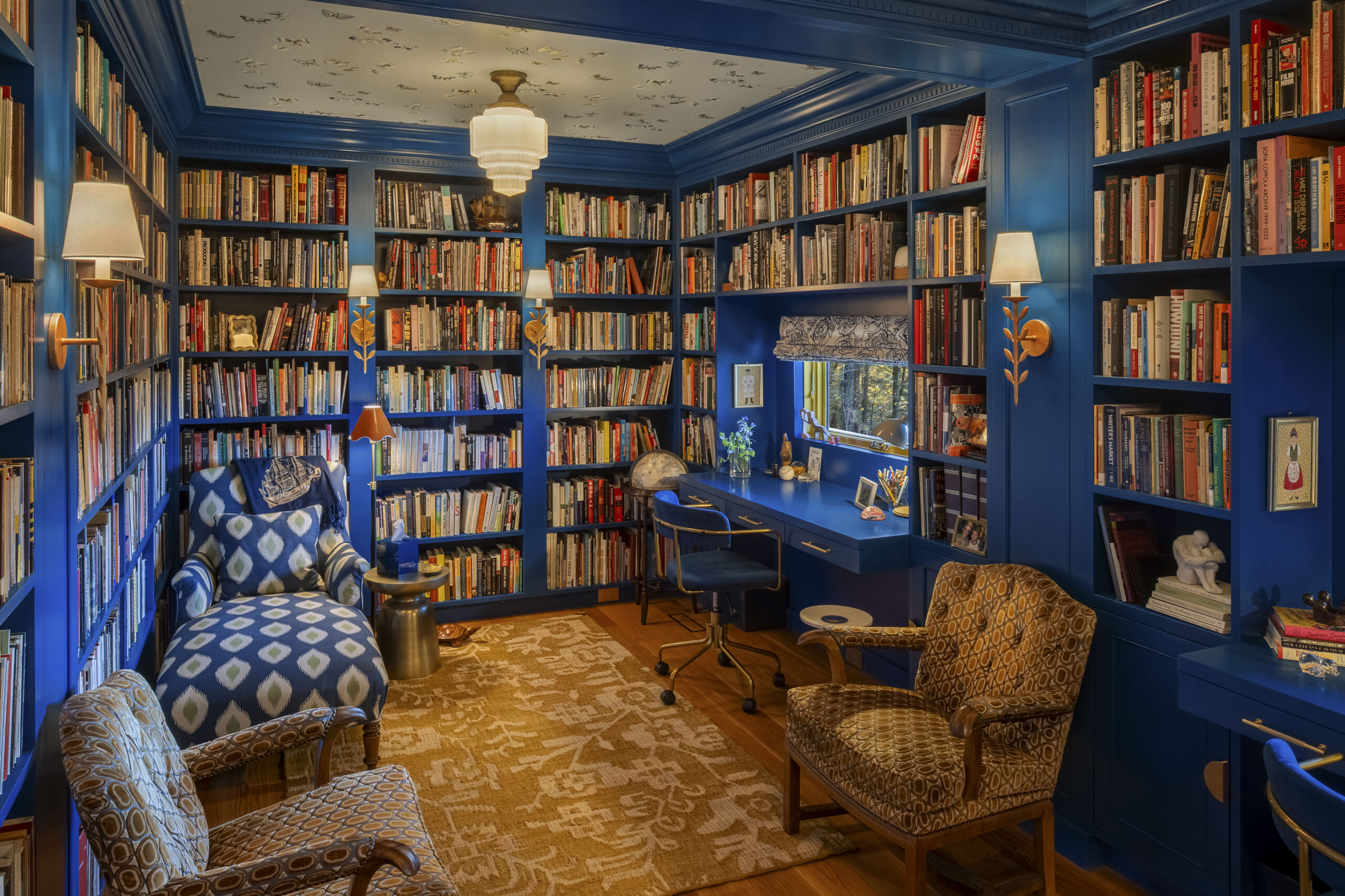 An alternate angle of the royal blue library, showing a cozy reading nook with a blue and white ikat-patterned chaise lounge. Gold-leaf sconces and an Art Deco ceiling fixture provide warm light, while a small window tucked into the bookshelves offers a glimpse of the autumn trees outside.