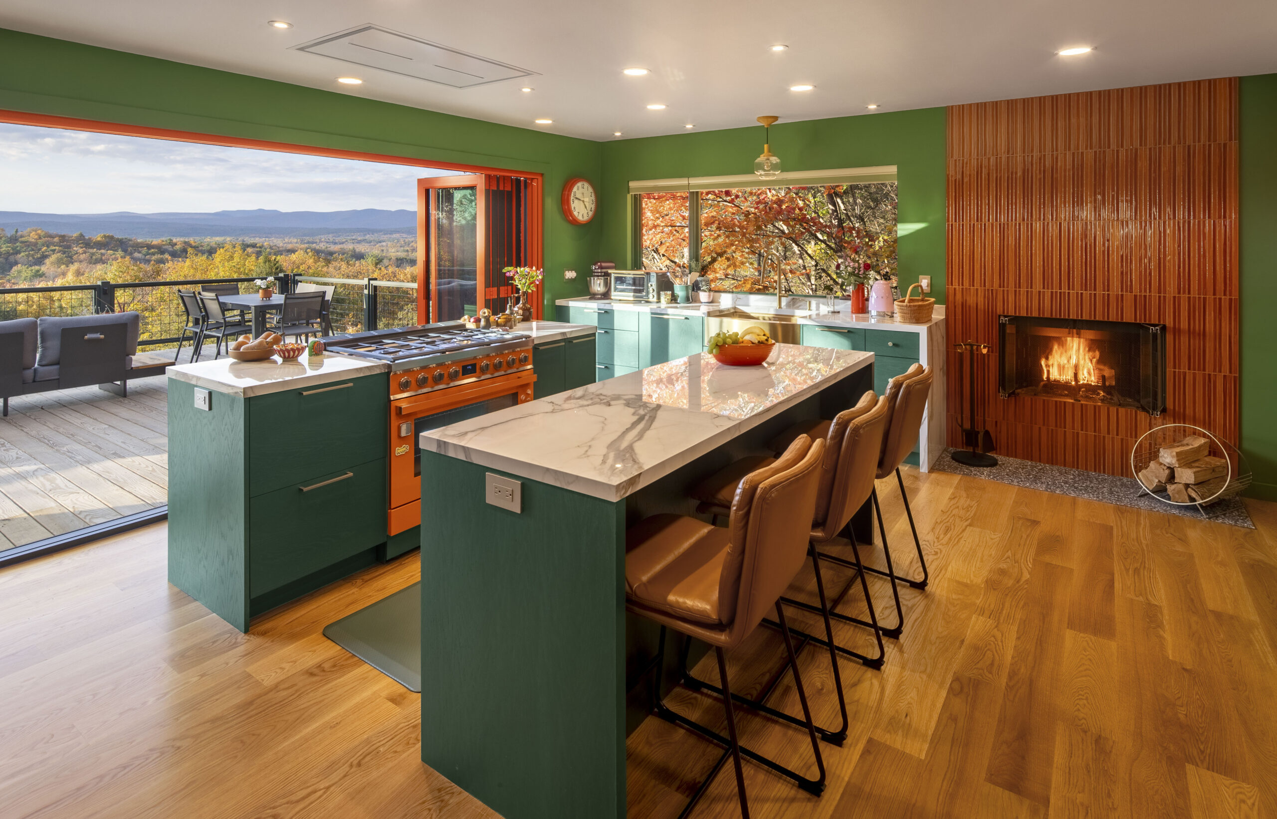 A perspective of the kitchen with the bifold doors wide open, showcasing an unobstructed view of the rolling mountains. The natural light highlights the contrast between the green walls, the marble island, and the warmth of the wood floors.