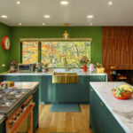 A close-up of the kitchen workspace featuring a large gold apron-front sink and brass fixtures. The green cabinetry and marble countertops are set against a window that perfectly frames the bright orange and yellow leaves of the forest outside.