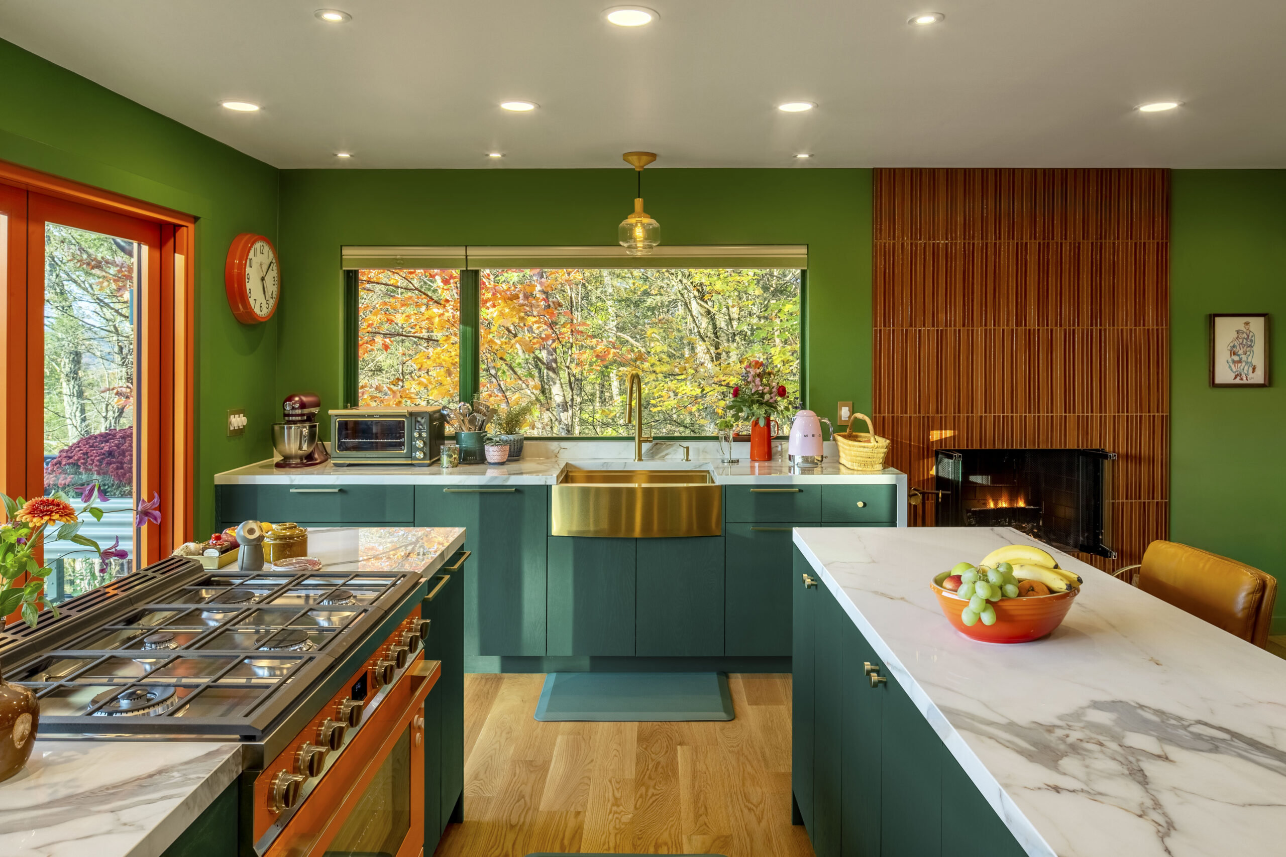 A close-up of the kitchen workspace featuring a large gold apron-front sink and brass fixtures. The green cabinetry and marble countertops are set against a window that perfectly frames the bright orange and yellow leaves of the forest outside.