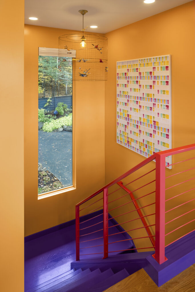 A vibrant stairwell featuring bright orange walls and a deep purple painted floor and staircase. A hot pink metal railing with horizontal cables adds a third layer of color. A whimsical birdcage-style chandelier with colorful bird figurines hangs above, while a vertical window offers a view of the Hudson Valley woods.