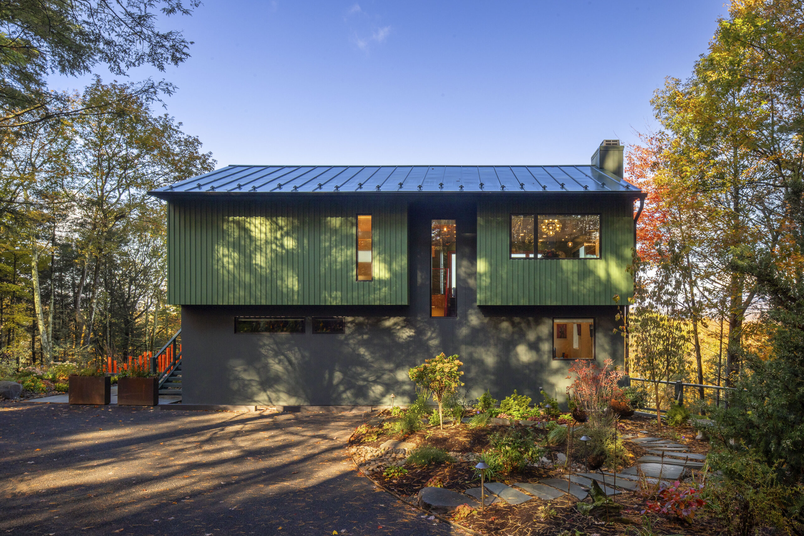A straight-on daytime view of the Woodland Pop House. The upper level is clad in vertical green siding, while the lower level is a smooth, dark grey. Minimalist landscaping with a winding stone path leads to the house, which is surrounded by lush Hudson Valley forest and dappled sunlight.