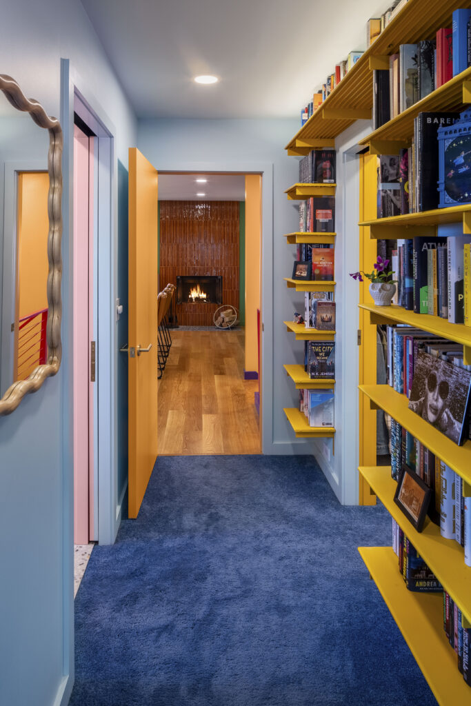A hallway with light blue walls and a plush deep blue carpet. An open orange door leads to a room with a glowing fireplace. On the right, bright yellow industrial-style shelves are packed with books, and a wavy-framed mirror on the left reflects the multi-colored doorways of the home.
