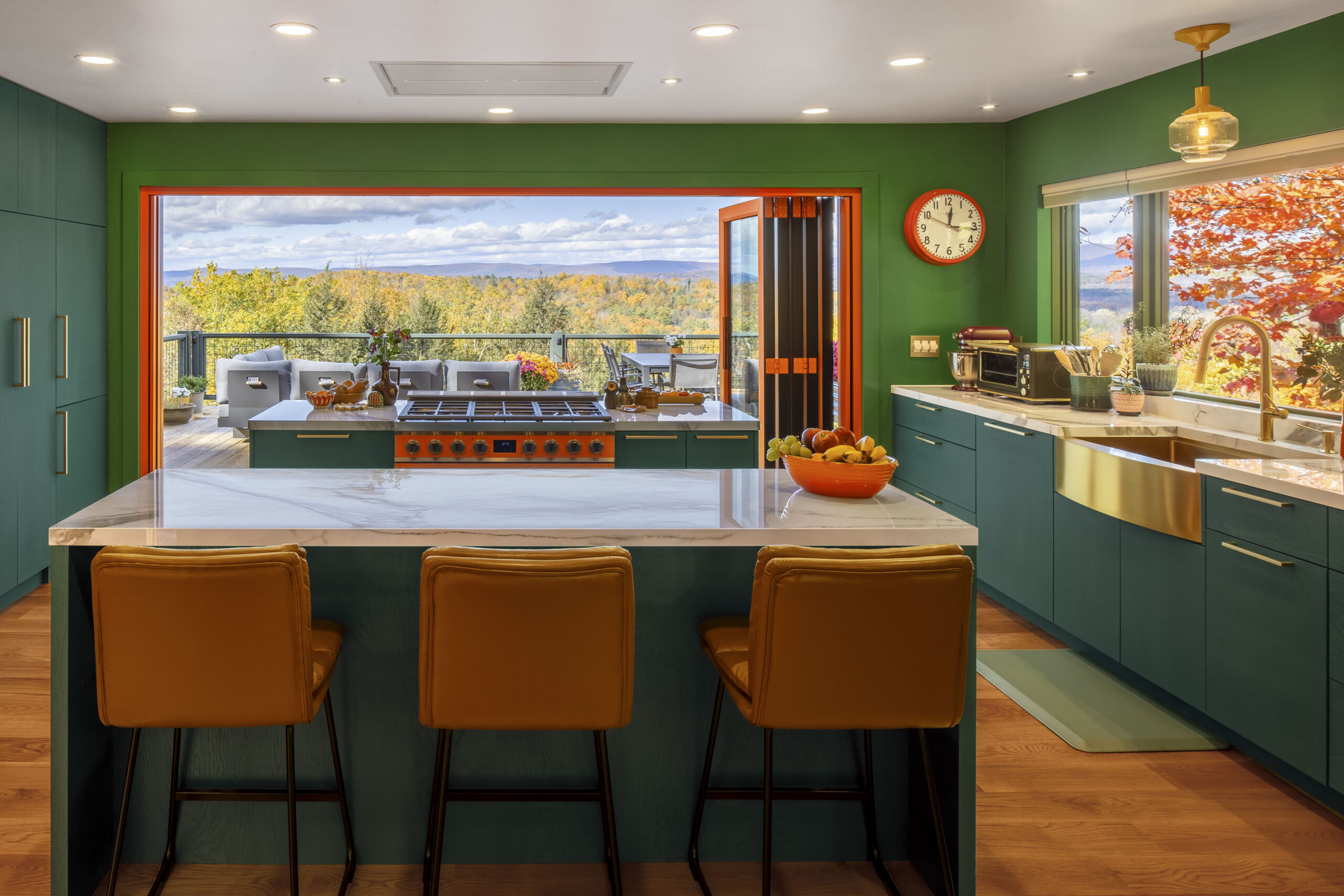 A view from behind the kitchen island looking toward the open deck. The tan leather barstools face the mountain vista. The room’s color palette—green, orange, and wood—creates a bridge between the modern interior and the natural Hudson Valley landscape.