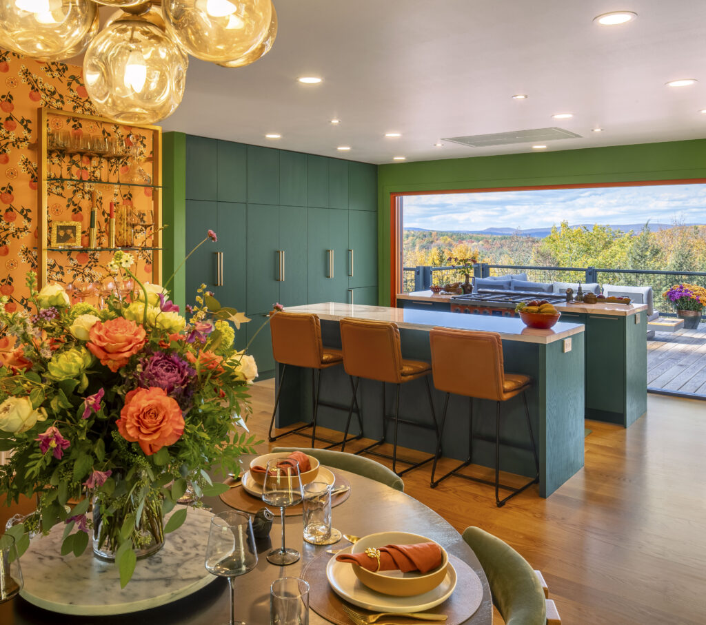 An interior view from the dining table looking toward the green kitchen. In the foreground, a large bouquet of orange and purple roses sits on a marble-topped table set with terracotta-colored dishes. Beyond the dining area, the forest-green cabinetry and the large open bifold doors create a seamless connection between the colorful interior and the Hudson Valley landscape.