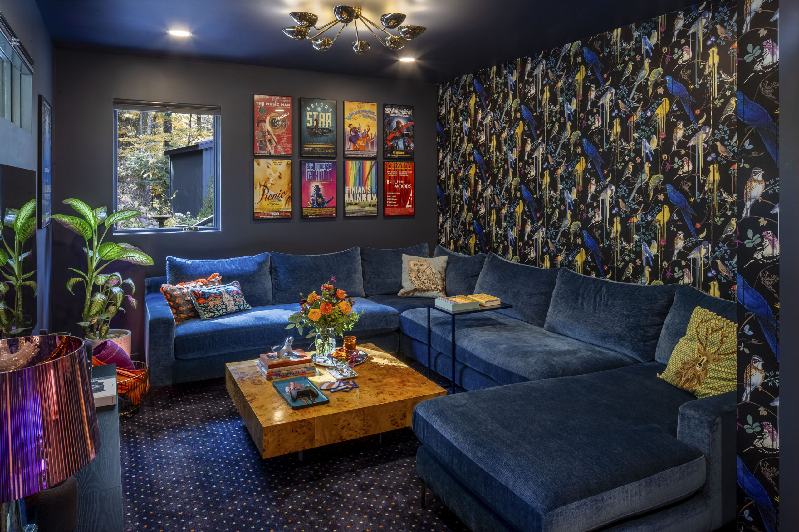 A cozy, dark-walled media room featuring a large deep blue sectional sofa. One wall is covered in a dark, illustrative bird-patterned wallpaper. A gallery wall of colorful vintage-style posters hangs above the sofa, and a large burl wood coffee table sits on a dark patterned carpet.