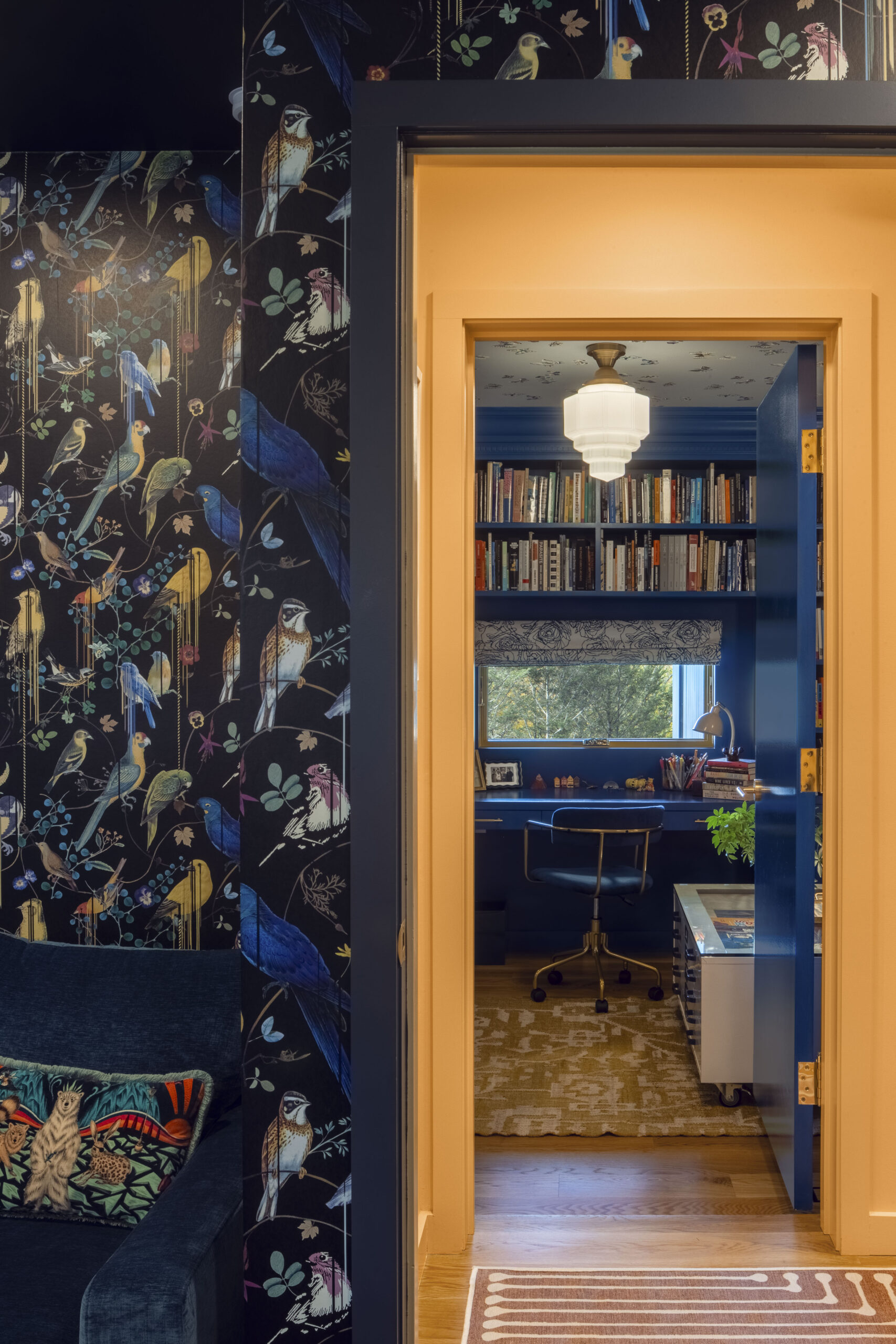A view looking through a bright orange door frame into a monochromatic blue library. The dark bird-pattern wallpaper of the foreground room contrasts sharply with the vibrant blue built-in bookshelves and desk visible in the next room, highlighted by an Art Deco-style pendant light.