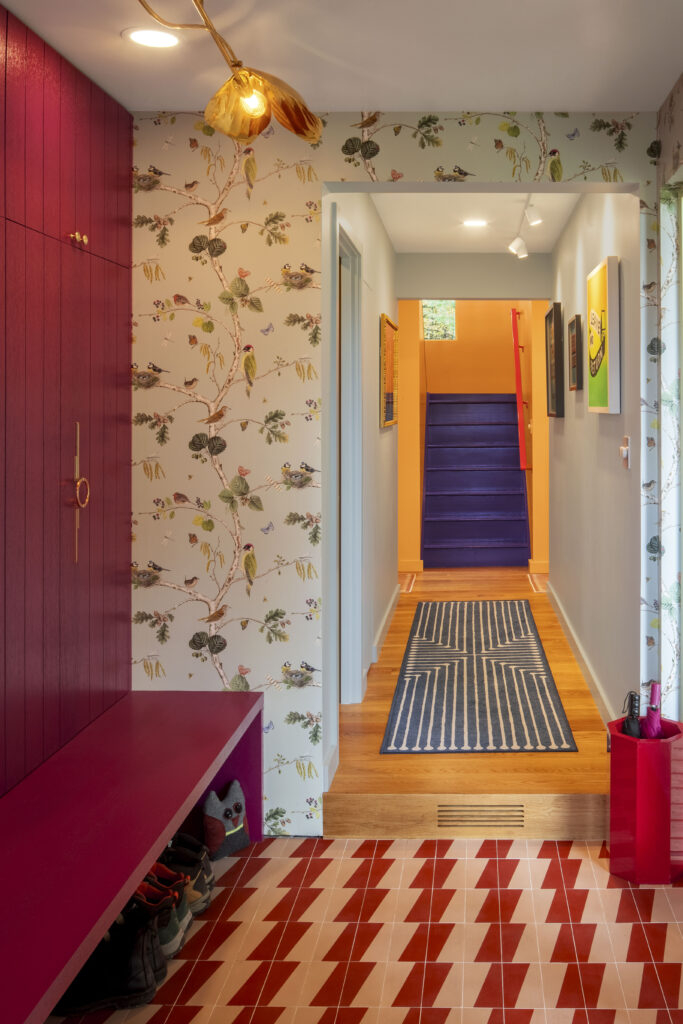 An entryway featuring a magenta-stained wood built-in bench and cabinetry against bird-and-branch patterned wallpaper. The floor is covered in red and white geometric tiles. A hallway with a blue geometric rug leads the eye toward the vibrant purple staircase and bright orange walls in the distance.