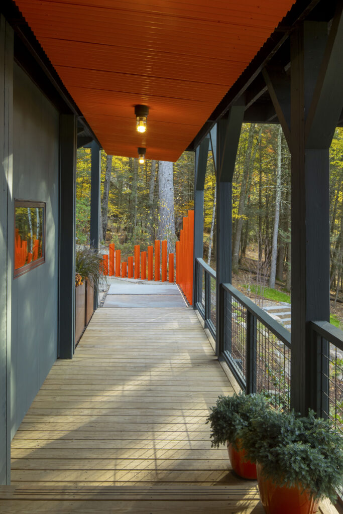 A first-person perspective looking down a wooden plank walkway toward a bright orange sculptural fence. The ceiling of the walkway is painted a bold, vibrant orange and is illuminated by industrial-style light fixtures. Potted evergreens in orange planters sit on the right, mirroring the home’s color accents.