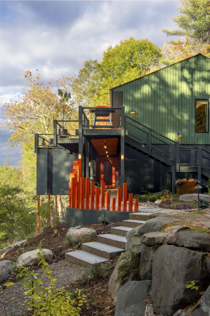 A low-angle shot of the house's entrance featuring large, natural stone steps leading up toward an orange-accented entryway. The bright orange vertical post fence creates a rhythmic barrier on the left, while the green siding of the upper floor provides a textural contrast against the soft, cloudy sky.